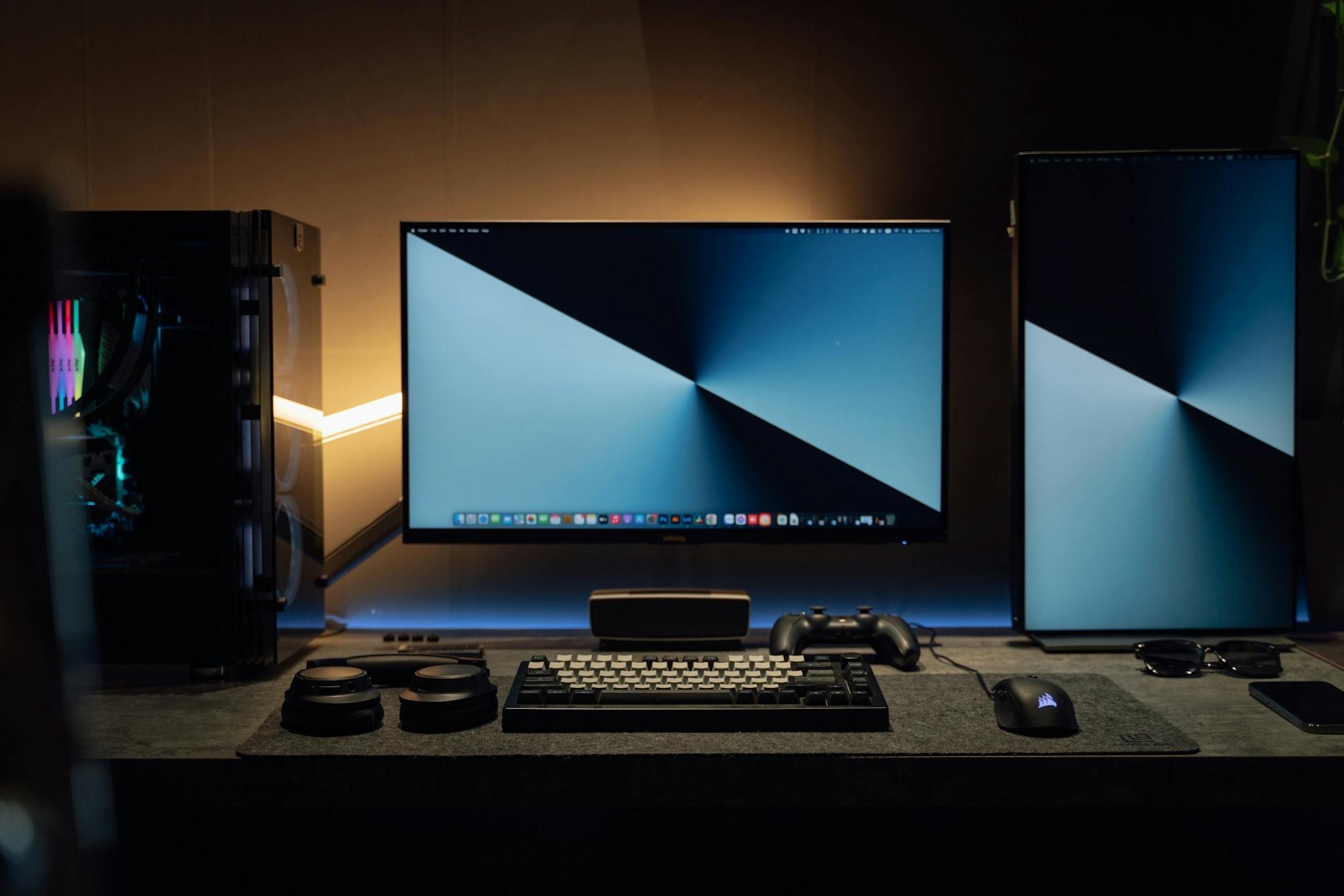 A dimly lit modern computer desk setup featuring dual monitors, a keyboard, and gaming accessories.